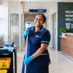 A smiling South African hospital cleaner holding a mop inside a bright, modern Gauteng Department of Health facility.