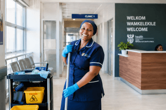 A smiling South African hospital cleaner holding a mop inside a bright, modern Gauteng Department of Health facility.
